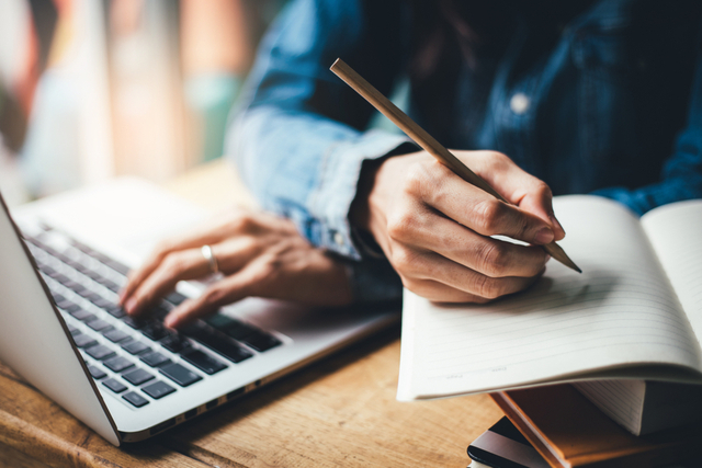 man writing in a notebook with one hand and typing on a computer with the other