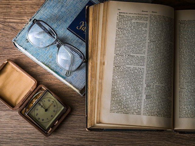 a dictionary, small carry clock and a pair of glasses on a wooden desk