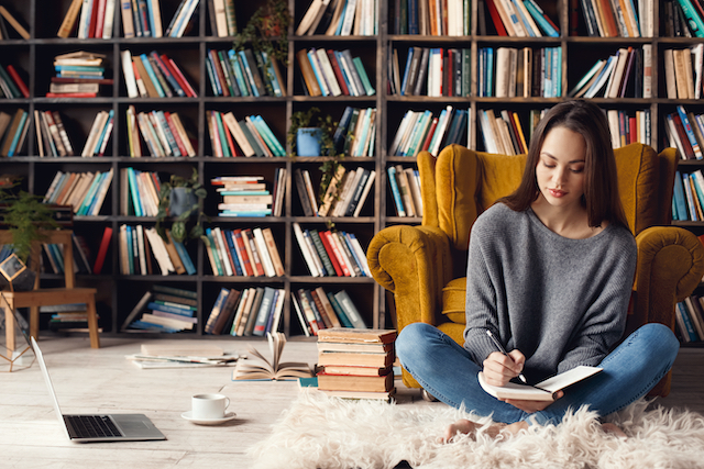 a woman sat on the floor writing in a notebook in front of some bookshelves