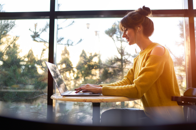 woman sat at a table by a window, typing on a laptop