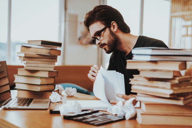 man sat at desk, writing, surrounded by crumpled paper and books