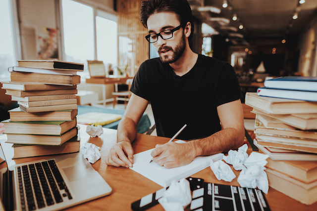 man writes at a desk piled high with books, a laptop, screwed up paper and a clapperboard