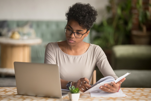 woman holding open book while looking at her computer