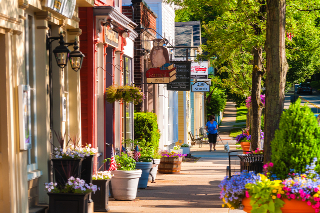 looking down a row of shopfronts. a woman walks a dog along the sidewalk.