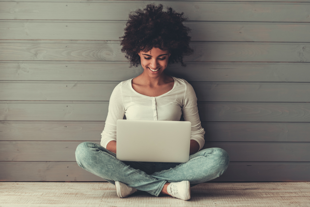 woman smiles while typing on laptop, sat against wall